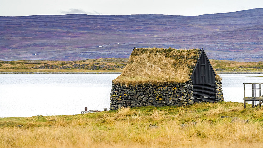 Stone building with grass roof facing the fjord, and a cross in the grass in front; Sudavik, Westfjords, Iceland