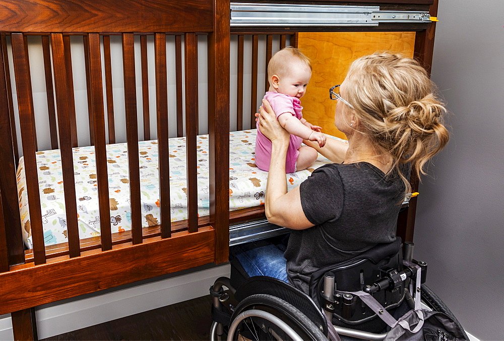 A paraplegic mother picking her baby up after a sleep in a customized crib with a sliding door; Edmonton, Alberta, Canada