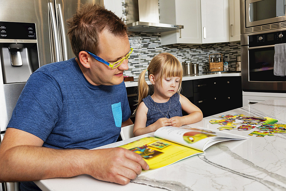 A father sitting down with his young daughter in the kitchen to read a book: Edmonton, Alberta, Canada