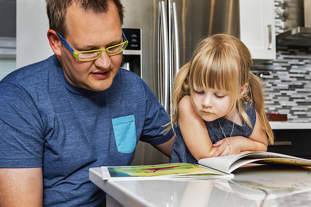 A father sitting down with his young daughter in the kitchen to read a book: Edmonton, Alberta, Canada