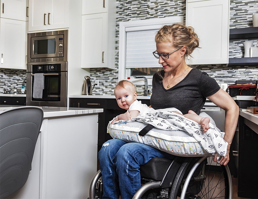 A paraplegic mother holding her baby on her lap, in her kitchen, while pushing in her wheel chair: Edmonton, Alberta, Canada