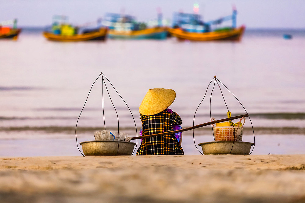 A woman sits on the beach looking out to the numerous fishing boats in the water off the coast, Ke Ga Cape; Ke Ga Island, Vietnam