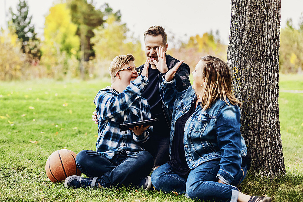 A young man with Down Syndrome celebrates with a high-5 after winning a game on his pad while enjoying quality time with his father and mother in a city park on a warm fall evening; Edmonton, Alberta, Canada