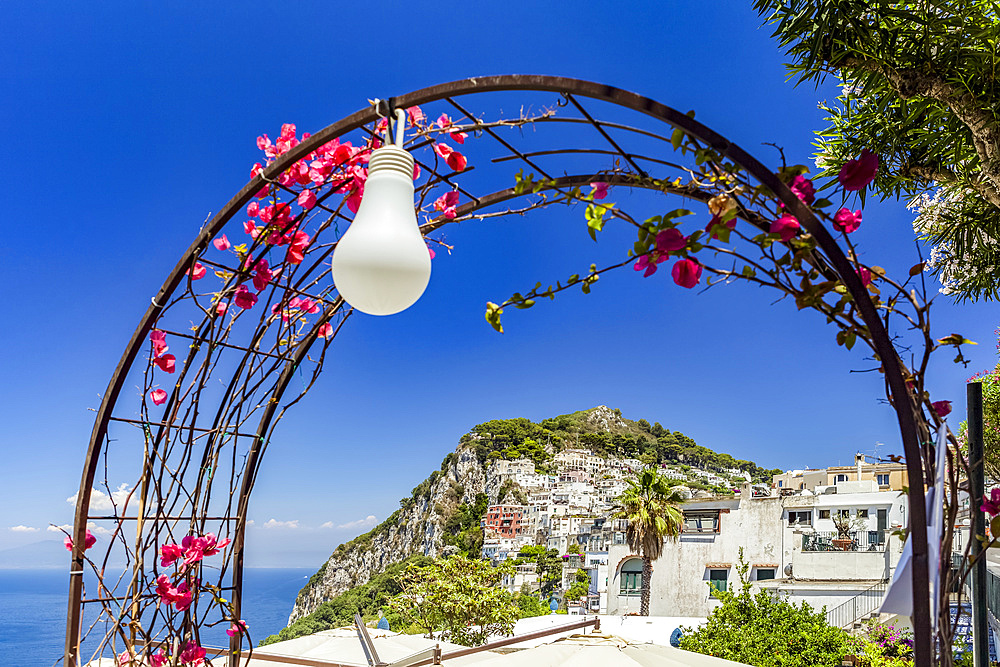 A decorative archway with light in the foreground and the city of Capri on the limestone crag along the coast of the Mediterranean; Capri, Italy