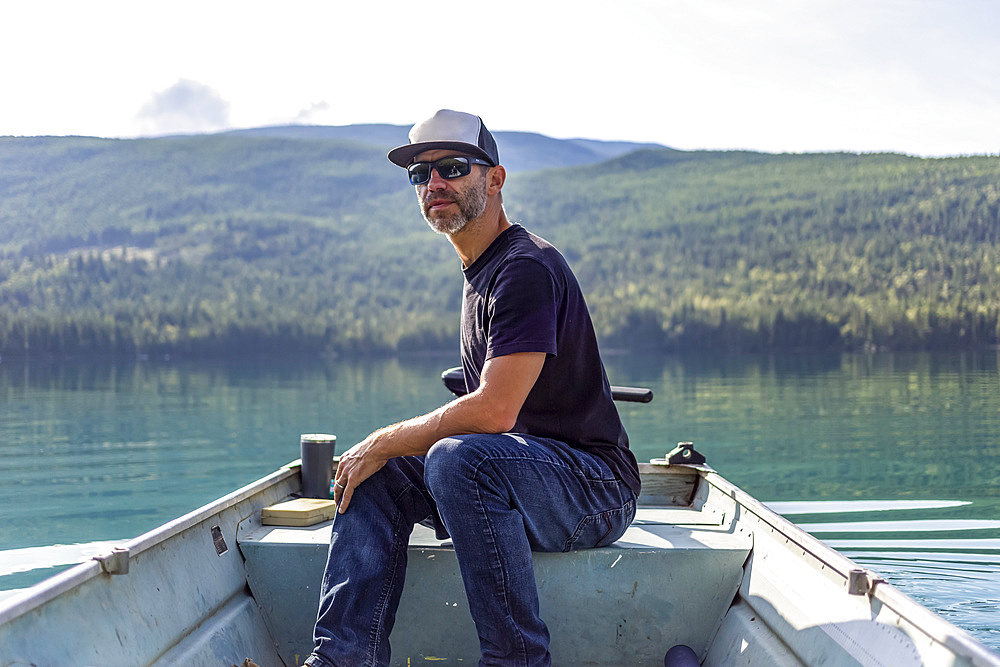 Man sits in a small rowboat with motor on White Lake with the tranquil water reflecting the forest in the background; British Columbia, Canada