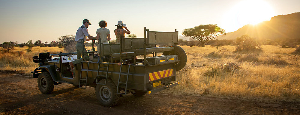 View taken from behind of a safari guide and women travelers standing in a parked jeep talking and looking out onto the savanna with binoculars on the Gabus Game Ranch at sunrise; Otavi, Otjozondjupa, Namibia