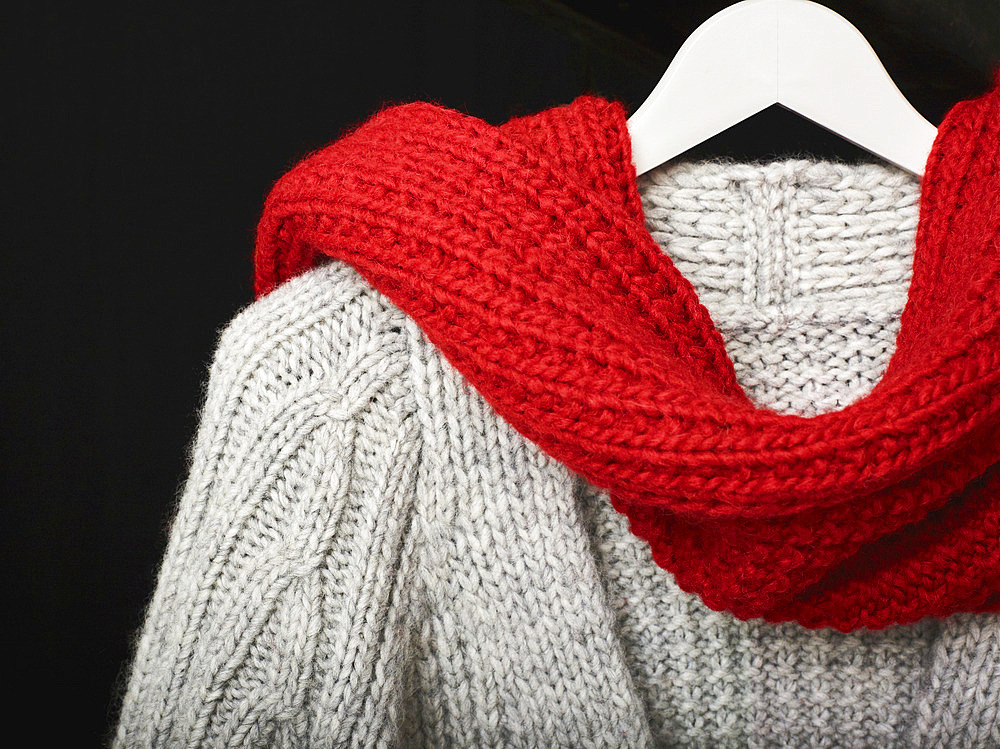 A grey knit sweater draped with a red knit scarf displayed on a hanger on a black background; Studio