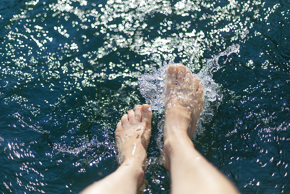 Looking down at bare feet splashing in water of Cultus Lake while sitting on a dock; British Columbia, Canada