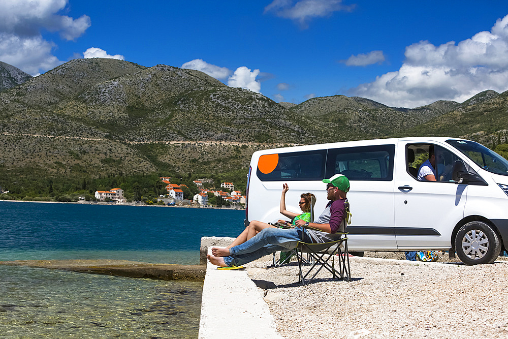 Friends sitting in the sun resting outside of a camper van as group of travelers stop in Slano for an afternoon at the beach; Slano, Dubrovnik-Neretva County (Dubrovačko-neretvanska županija), Croatia