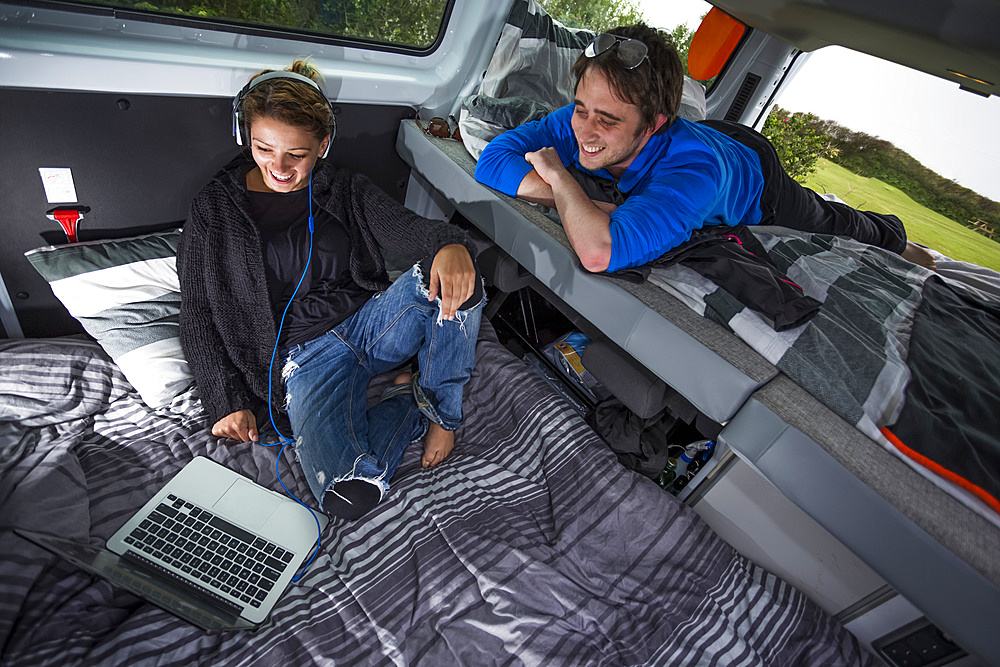 The inside of a campervan becomes a work station and living room. A young couple relaxing while watching the screen of a laptop computer; Penzance, England, United Kingdom