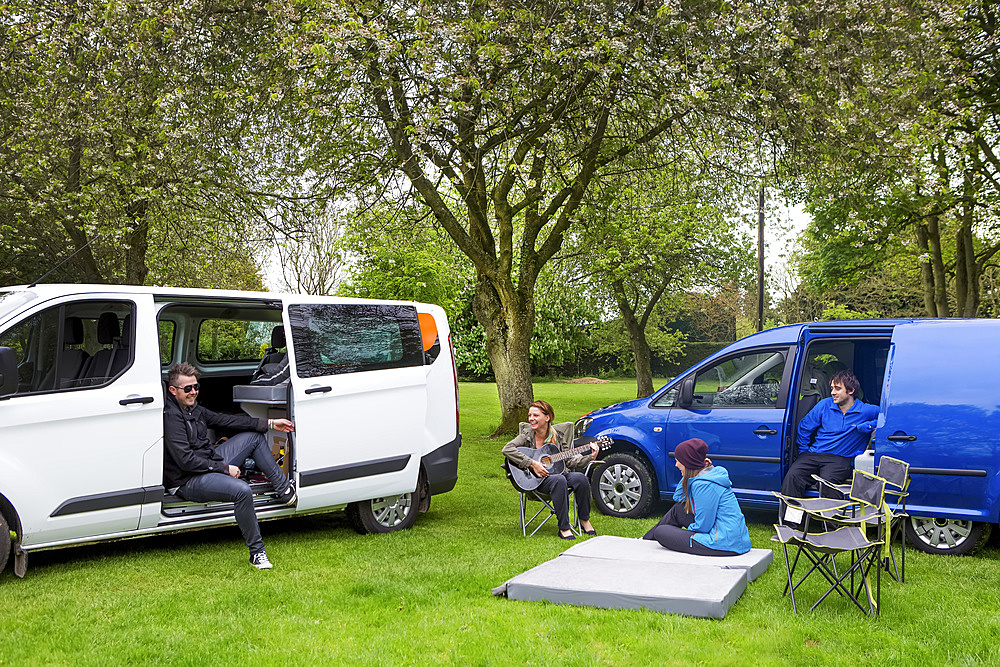 Friends relaxing while setting up camp outside two modified caravan campers; Bourton-on-the-Water, England, United Kingdom