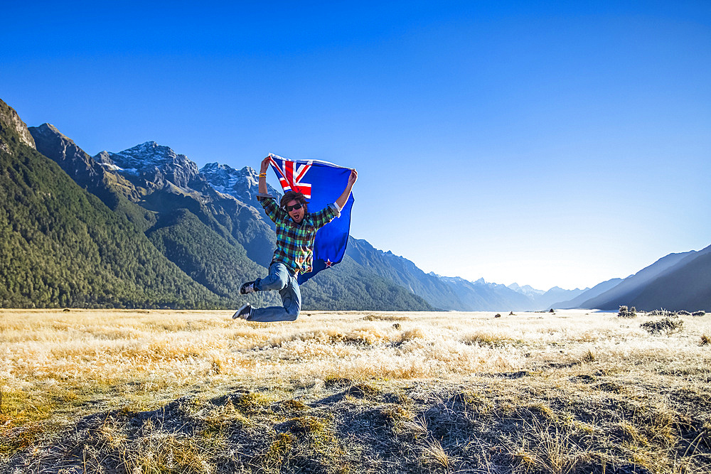 A man jumps in the air with the New Zealand flag in Fiordland National Park; Southland, New Zealand