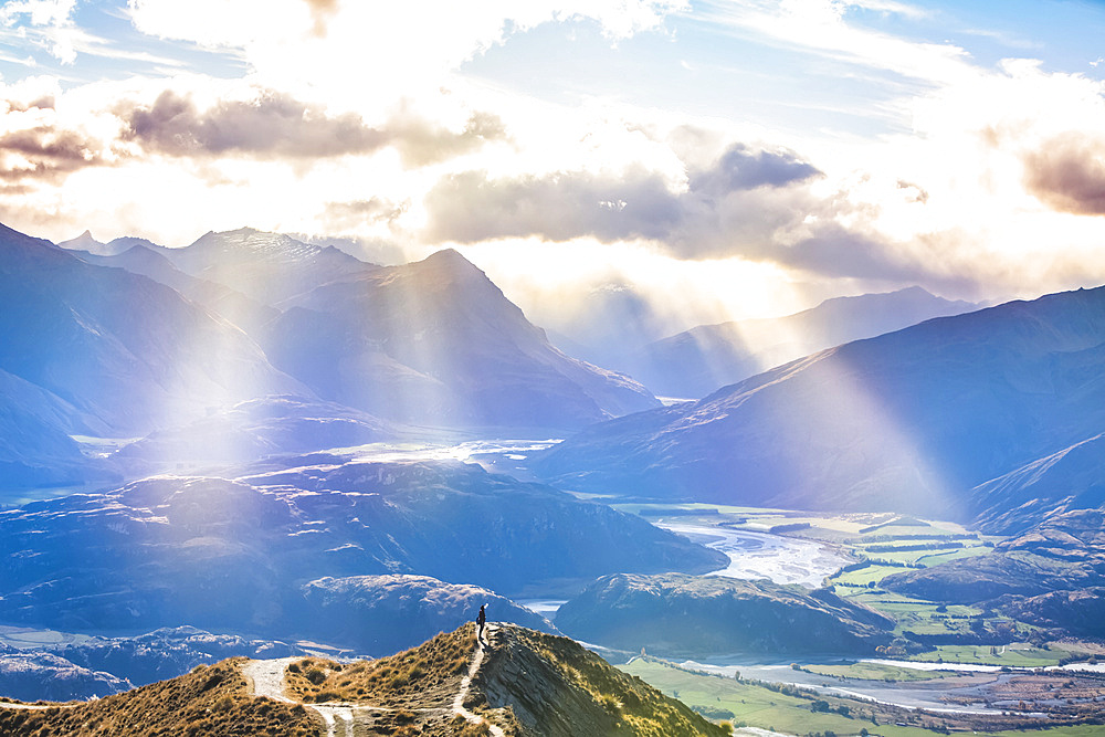 Man standing on mountaintop at Roys Peak with beautiful rays of sunlight after the strenuous hike to the lookout to see the spectacular views of the lakes and surrounding mountains of the Southern Alps near Wanaka; Otago, New Zealand