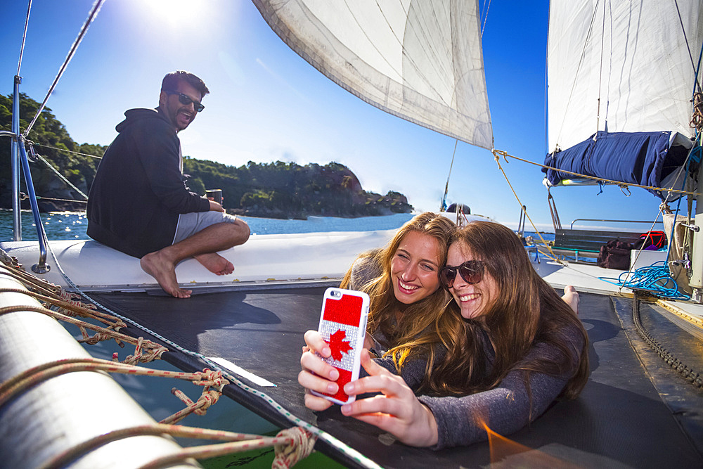 Friends taking a selfie onboard a catamaran boat tour through Abel Tasman National Park with a man smiling in the background; Tasman, New Zealand