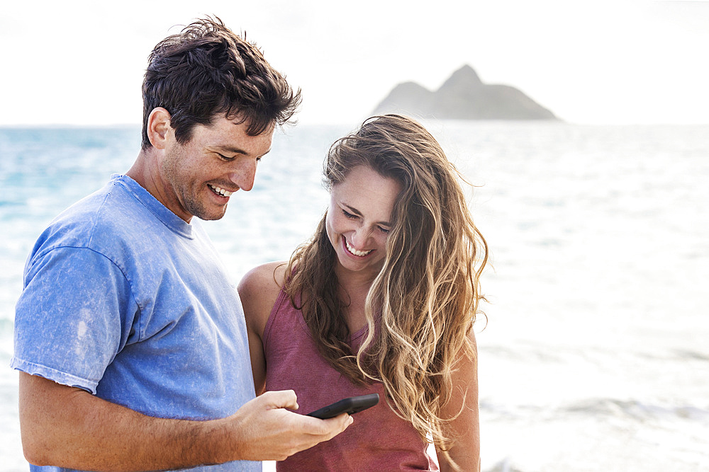 A young Millennial couple looking at a smart phone on Lanakai Beach with the Mokes Islands in the background; Lanakai, Oahu, Hawaii, USA