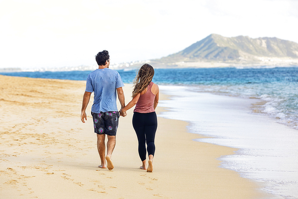 A view from behind of a Millennial couple walking along Lanikai Beach with Mokapu Point in the background; Lanikai, Oahu, Hawaii, USA