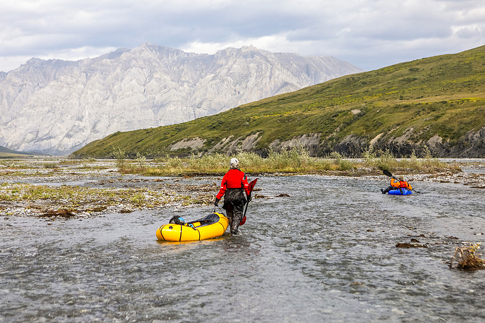 Caucasian woman wearing red drysuit, drags her yellow packraft down a shallow braid of the Marsh Fork river, while a caucasian man wearing orange drysuit paddles a blue boat downriver, through the Brooks Range mountains, Arctic National Wildlife Refuge, Alaska on a sunny summer day; Alaska, United States of America