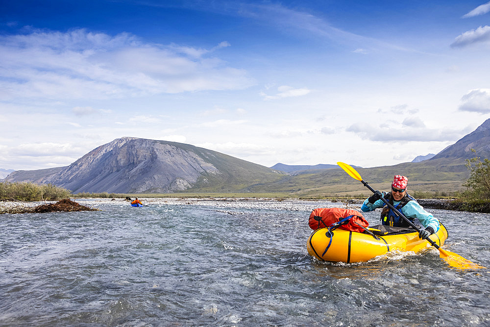 Caucasian woman in her 40's smiles as she digs deep with her paddle into the river, guiding her yellow packraft down the crystal clear Marsh Fork River, in the Brooks Range, on a sunny summer day, Arctic National Wildlife Refuge, Alaska, while another boat is in the background way behind her; Alaska, United States of America