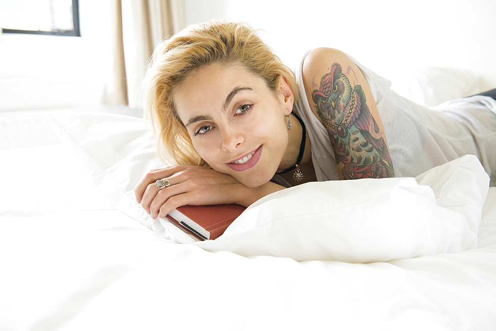 A young woman with tattoos lying on a bed with a book.