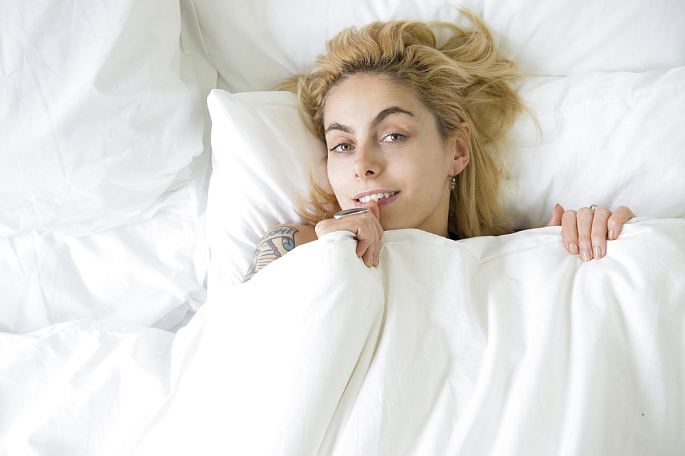 A cool young woman lying in a bed with he covers pulled up to her chin.