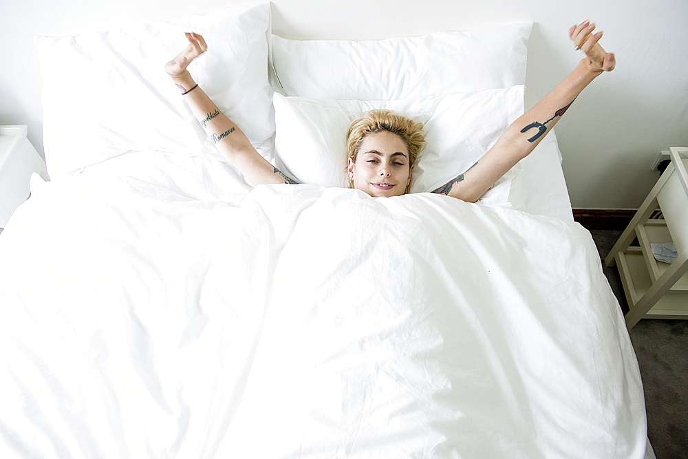 A cool young woman lying in a bed stretching her arms.