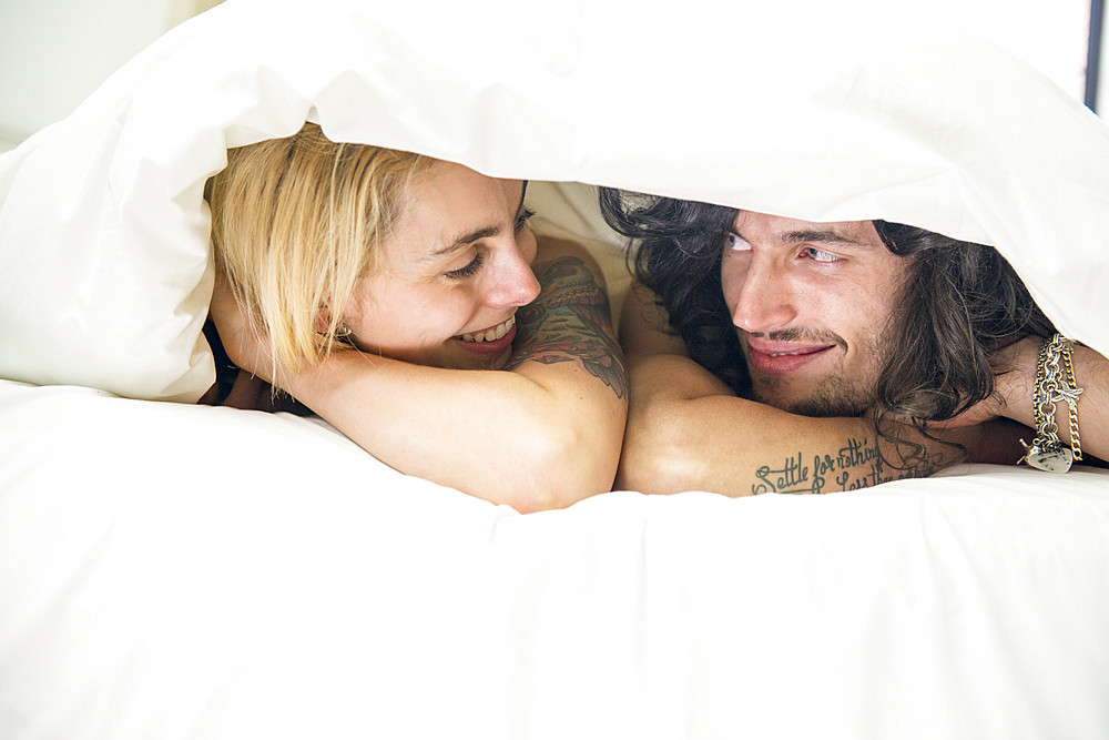 A cool young tattooed couple peeking out from under a duvet on a bed.