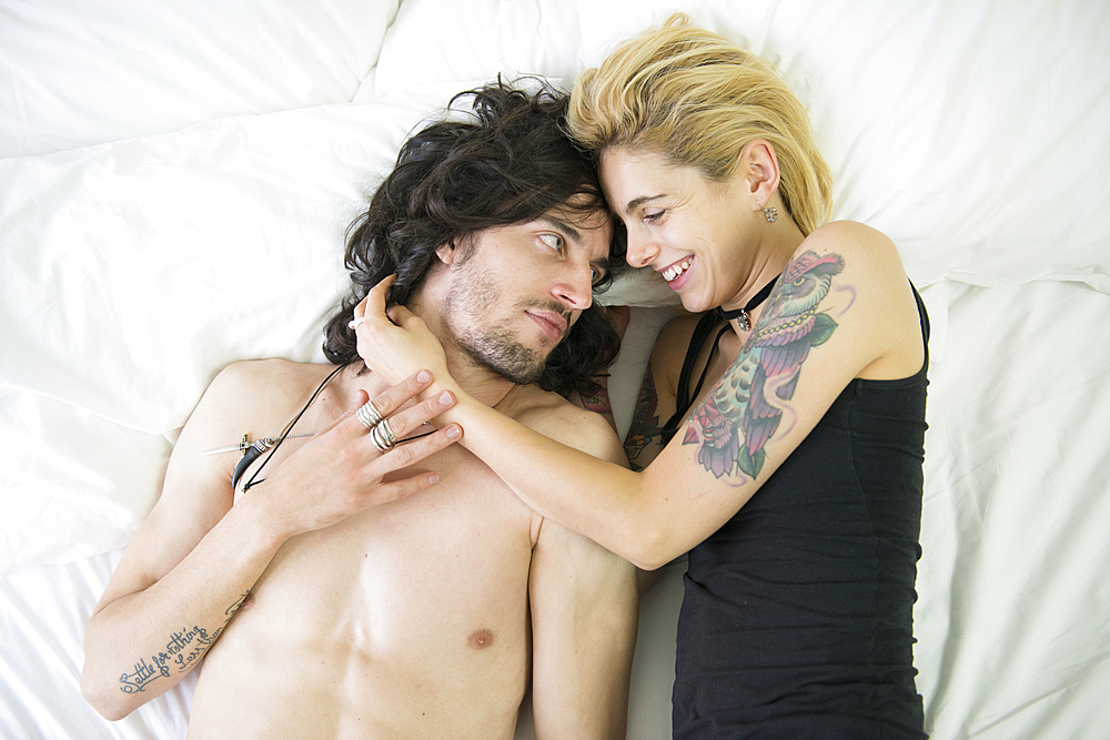 A cool young tattooed couple cuddling on a bed.