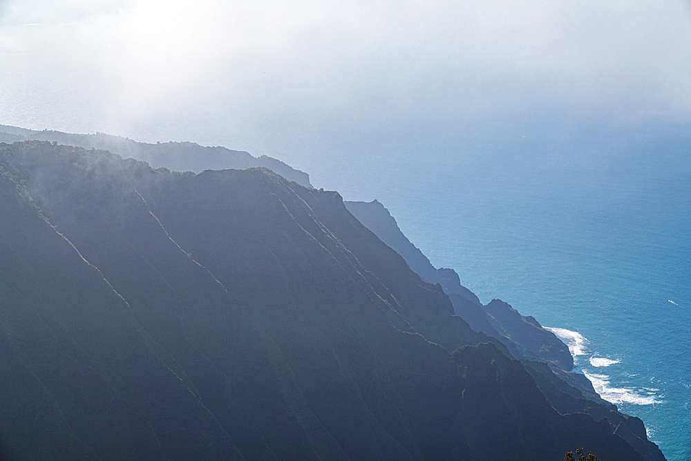 Hazy view of the steep, silhouetted mountain cliffs of the Napali Coast along the Kalalau Trail on the Hawaiian Island of Kauai against the blue waters of the Pacific Ocean, Kauai, Hawaii, United States of America