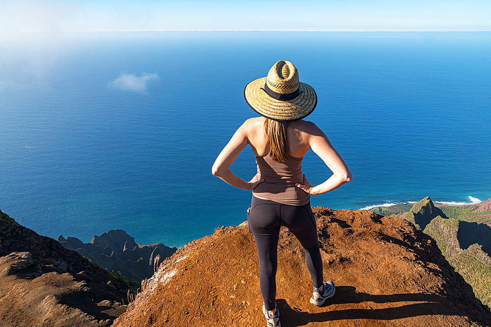 View taken from behind of a woman in a straw hat standing on the top of a mountain cliff looking out onto the brilliant, blue water of the Pacific Ocean along the Kalalau Trail on the Napali Coast, Kauai, Hawaii, United States of America