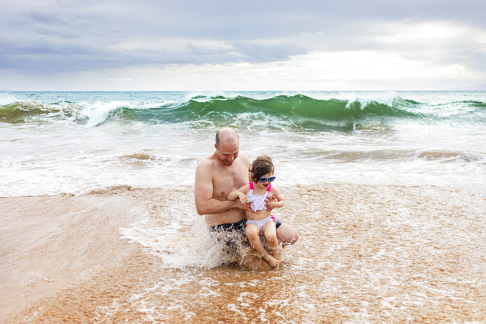 Father and daughter sitting in the water on the shoreline playing in the foamy surf along Kamaole Beach, Maui, Hawaii, United States of America