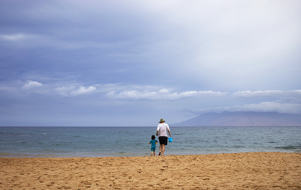 Scenic view, taken from behind, of a grandmother and granddaughter walking along the shore at Maluaka Beach with the Pacific Ocean and cloud covered mountains in the distance, Maui, Hawaii, United States of America