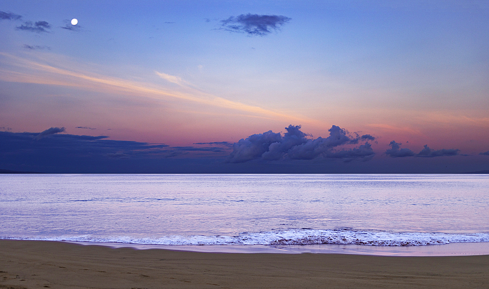 Ocean surf breaking on the shore along Kamaole 2 Beach at twilight with the moon high in the blue sky with a pink hue over the clouds and reflected on the water of the Pacific, Kihei, Maui, Hawaii, United States of America