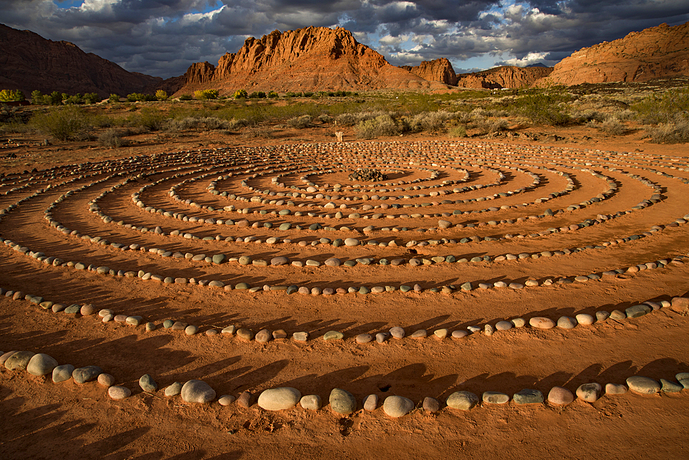 Hiking trail through Snow Canyon, with circles of stones in a meeting area behind the Red Mountain Spa, with meditation maze and Snow Canyon Mountain Range in the background. Red Cliffs Desert Reserve around St George Town with rock cliffs and dark clouds in a blue sky, St George, Utah, United States of America