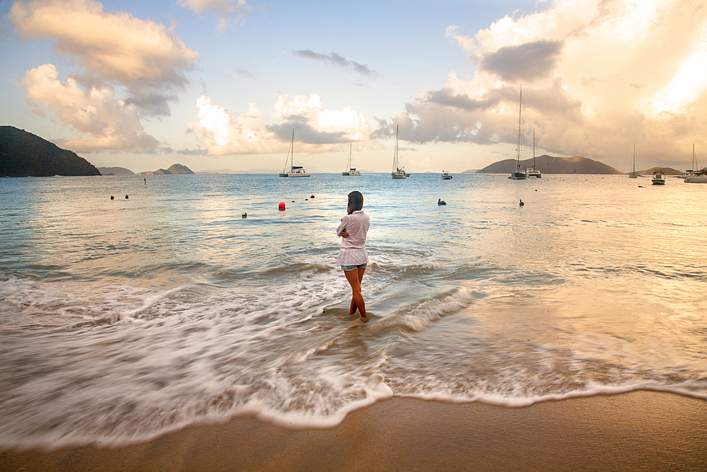 View taken from behind of a woman standing in the foamy surf on the beach, looking out at the turquoise water with boats moored off shore along the horizon in Cane Garden Bay at twilight, Tortola, British Virgin Islands, Caribbean