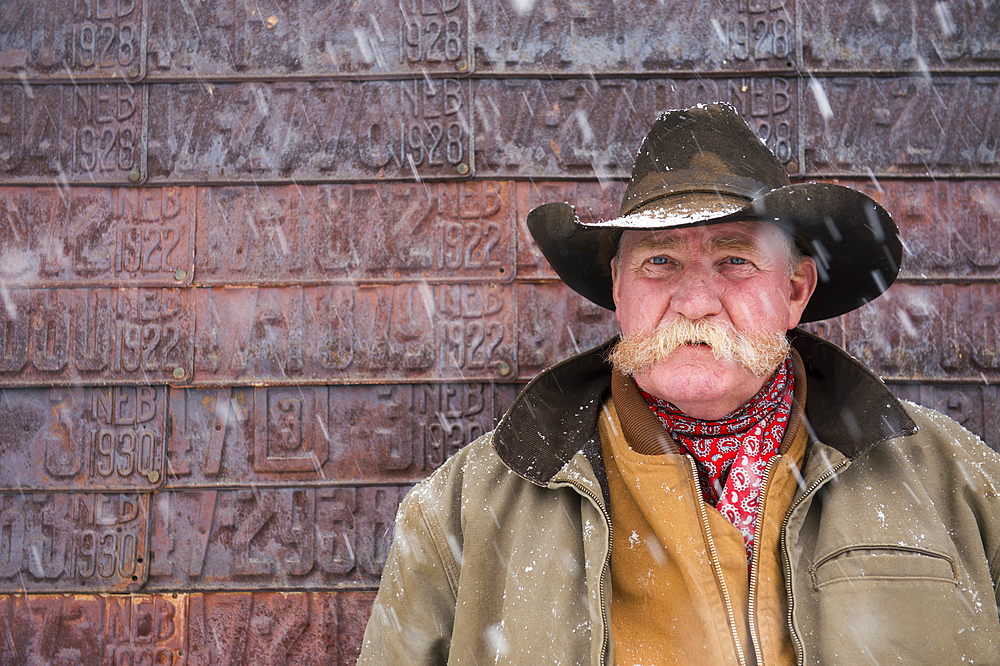 Portrait of a man in a cowboy hat standing by a barn wall covered with old license plates during a snow storm, Burwell, Nebraska, United States of America