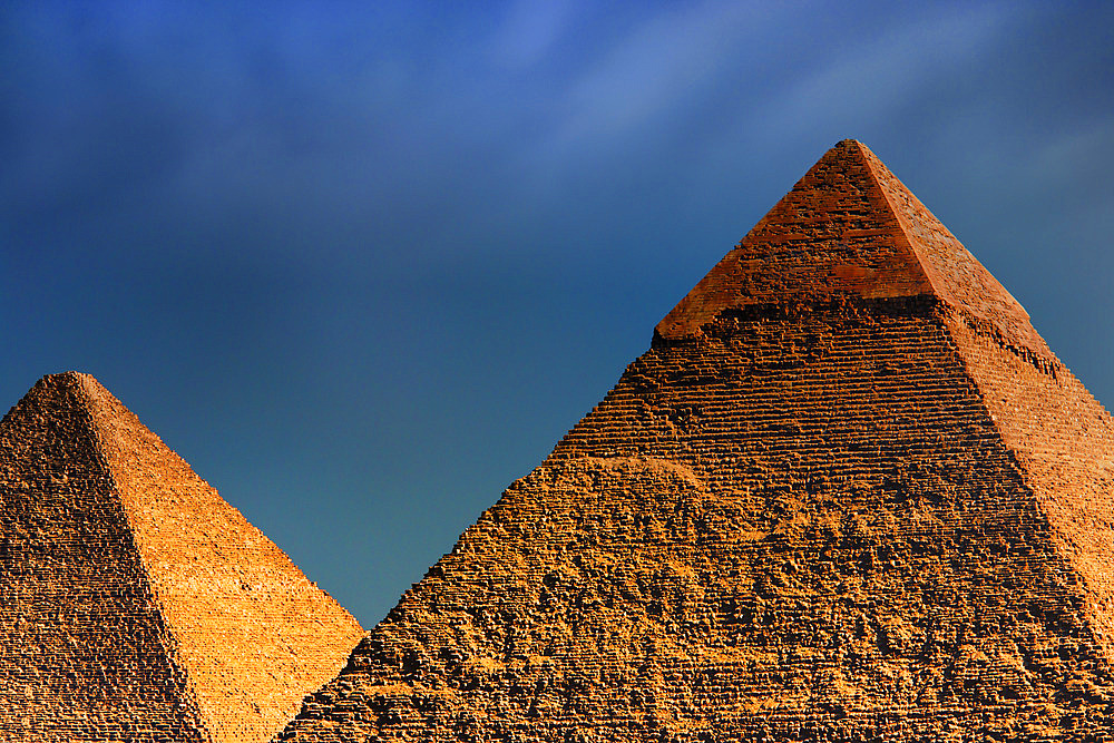 Two of the primary Pyramids of Giza, sunlit with a golden hue, the Great Pyramid (Khufu) under a dark, cloudy sky, Giza, Egypt