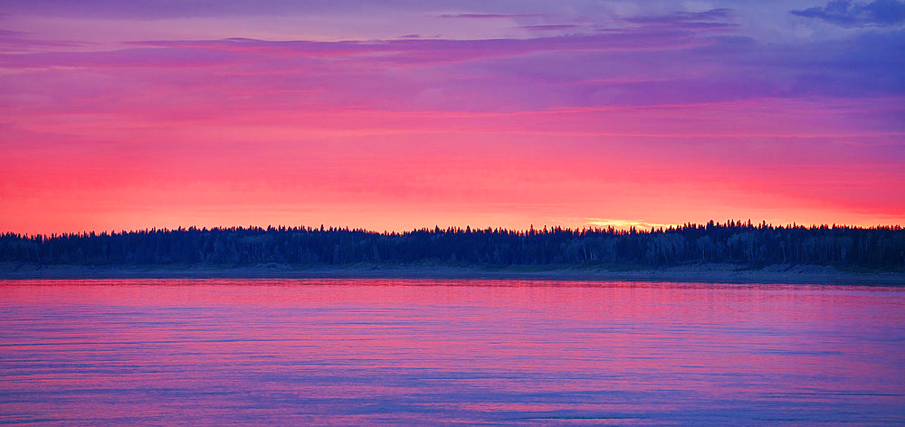 The evening sky ignites with colour as the sun goes below the horizon line in Fort Simpson. The Mackenzie River reflects the colour in a beautiful moment, Fort Simpson, Northwest Territories, Canada