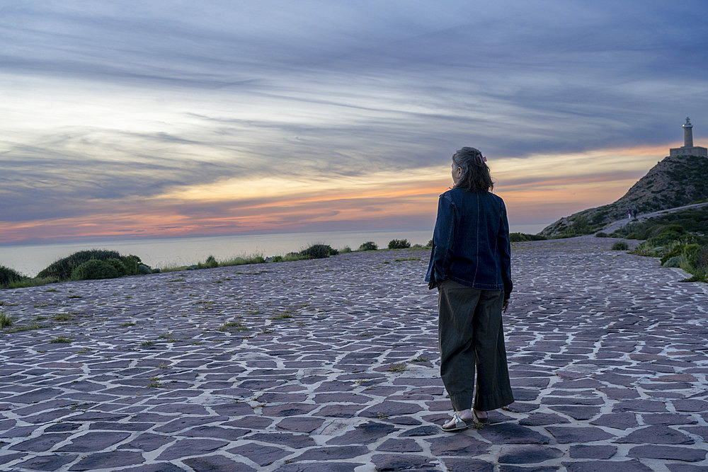 Woman in a long coat stands looking out over the Mediterranean Sea at sunset, with Capo Sandalo Lighthouse in the background, on San Pietro Island on the southwestern coast of Sardinia, Italy, Carloforte, South Sardinia, Italy