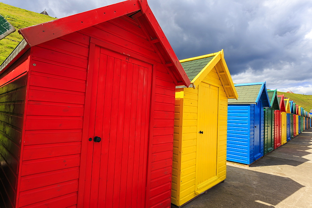 Row of colourful beach huts and their shadows, with grassy cliffs, West Cliff Beach, Whitby, North Yorkshire, England, United Kingdom, Europe