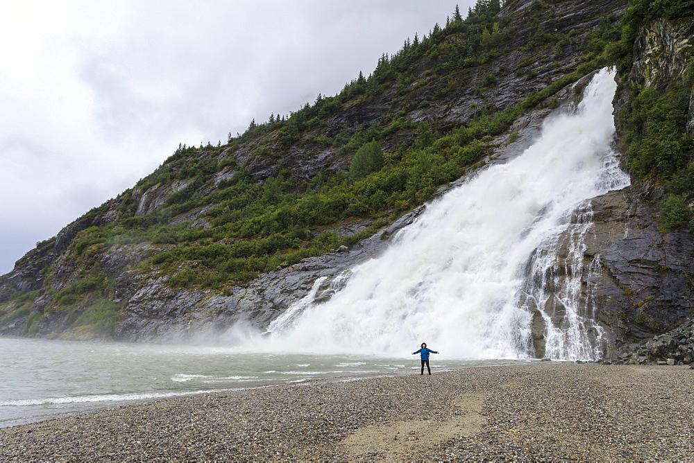 Visitor with outstretched arms on a beach in front of Nugget Falls Cascade, Mendenhall Lake and Glacier, Juneau, Alaska, United States of America, North America