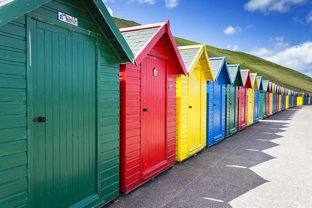 Row of colourful beach huts and their shadows with green hill backdrop, West Cliff Beach, Whitby, North Yorkshire, England, United Kingdom, Europe