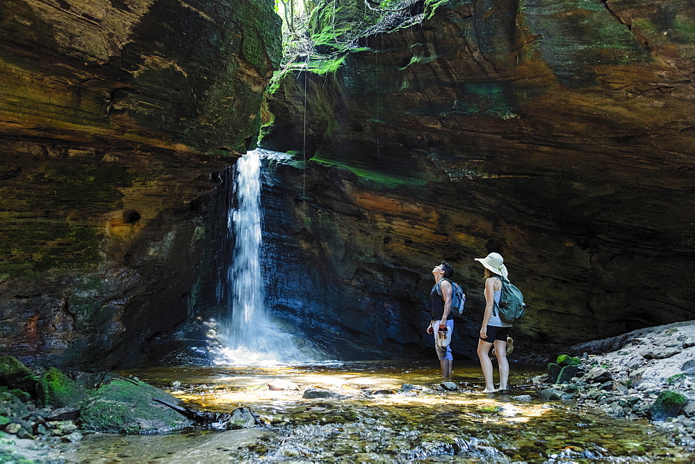 Two young hikers canyoning up a river and standing in a cave in the rainforest, in front of a pristine waterfall, Minas Gerais, Brazil, South America