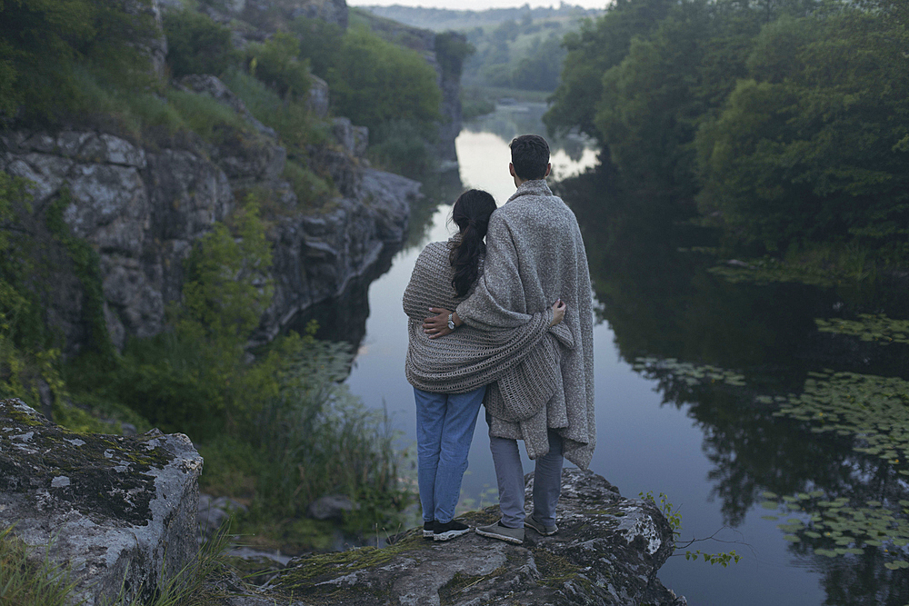 Caucasian couple wrapped in blanket standing on rock admiring river