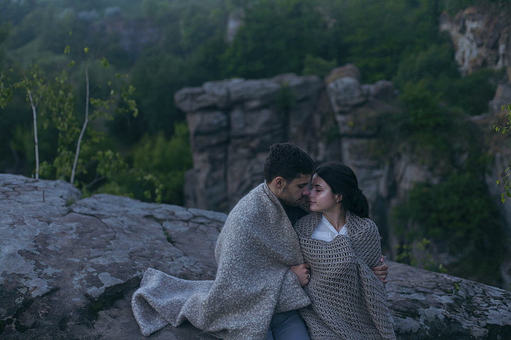 Caucasian couple sitting on rock wrapped in blanket