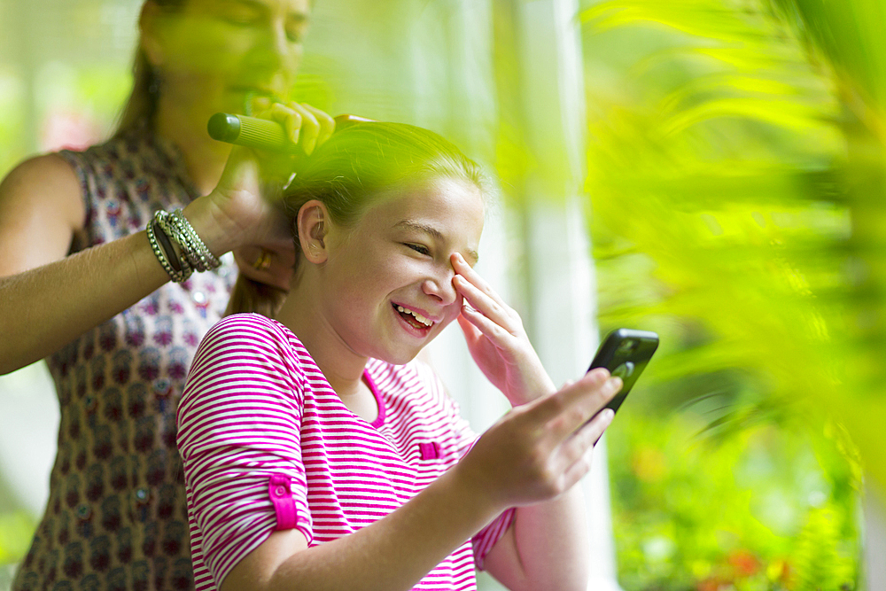 Caucasian mother brushing hair of daughter texting on cell phone