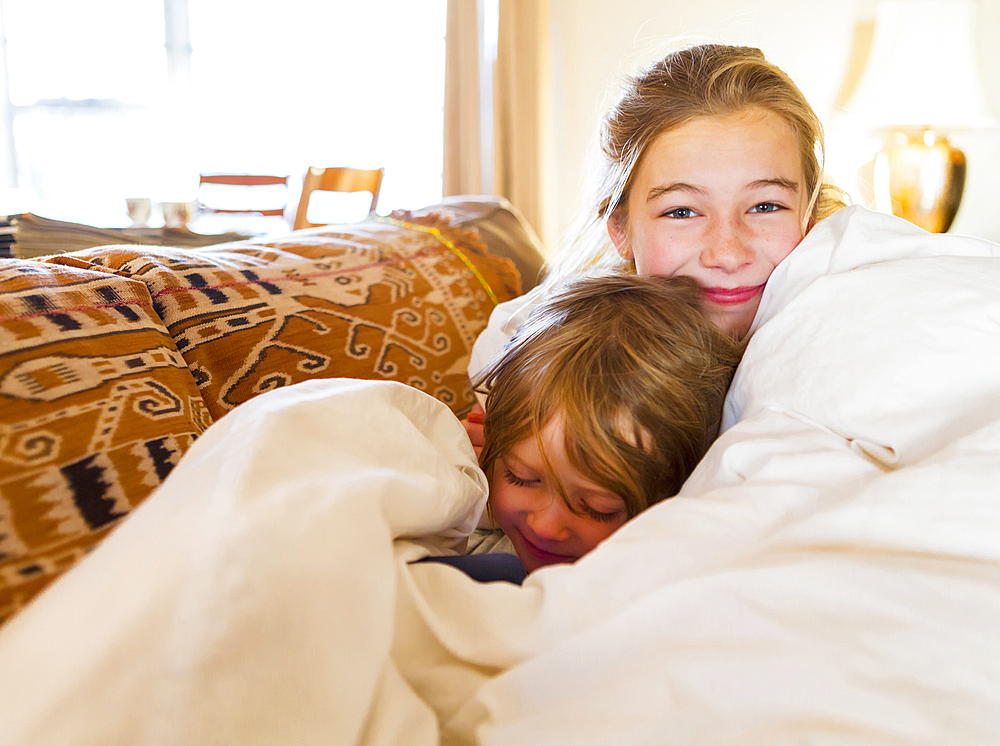 Caucasian brother and sister cuddling in comforter