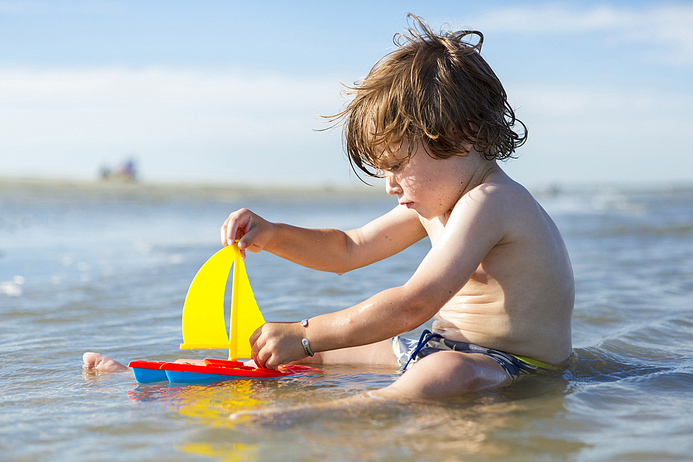 Caucasian boy sitting in ocean playing with toy sailboat