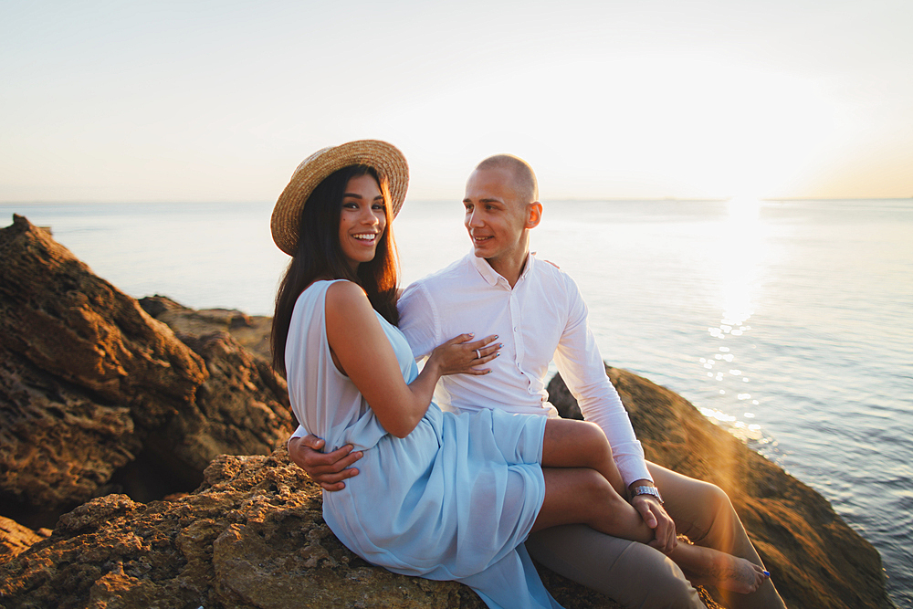 Caucasian couple sitting on rocks on beach at sunset