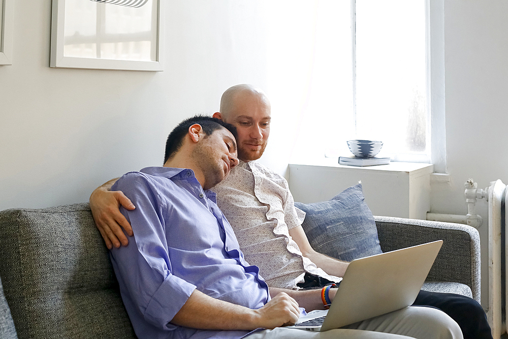 Caucasian men hugging on sofa and using laptop