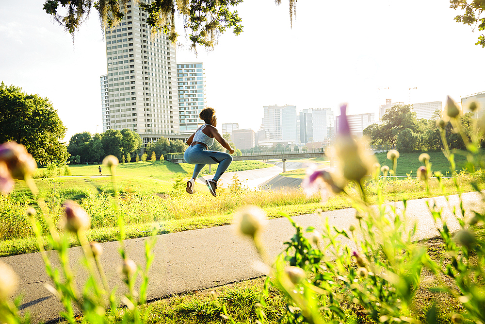 Mixed Race woman jumping on running path in park beyond wildflowers